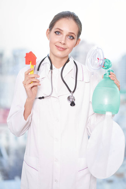Une jeune femme médecin se tient debout dans un blouse blanche, regardant directement dans la caméra, tenant une petite maison de jouets et un ventilateur pulmonaire artificiel. Soins de santé, quarantaine, pandémie
                                - Photo, image