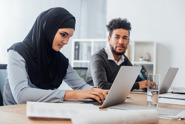 Селективный фокус арабской предпринимательницы с помощью ноутбука рядом с африканским американским коллегой в офисе
  - Фото, изображение