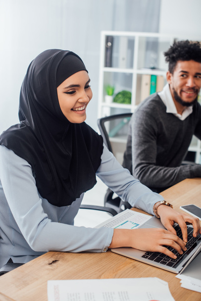 Selective focus of smiling arabian businesswoman using laptop near african american colleague in office  - 写真・画像