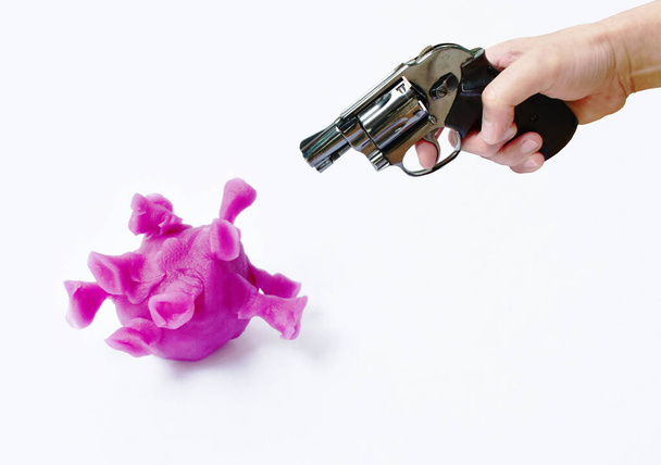 Hand hält Revolver Pistole vorbereiten, um lila Coronavirus oder COVID-19 Skulptur aus Ton auf weißem Hintergrund zu schießen  - Foto, Bild