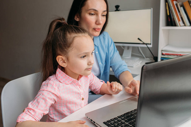 Anya és lánya ma otthon vannak. Távolról dolgoznak egy laptopon. Anya segít a lányának befejezni a feladatokat a számítógépen. - Fotó, kép