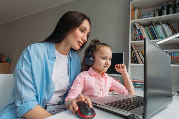 Mamá y su hija están en casa hoy. Trabajan remotamente en un portátil. Mamá ayuda a su hija a completar tareas en una computadora
. - Foto, imagen