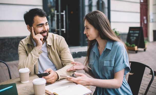 Cheerful male and female friends discussing new smartphone notification during knowledge time in street coffee shop, happy couple in love talking about ideas for joing blog in social networks - 写真・画像