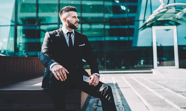 Contemplative male corporate employee sitting on urban setting and thoughtful looking away pondering on idea for startup project with executive managers, pensive businessman in financial district - 写真・画像