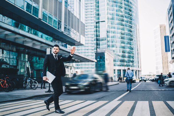 Caucasian confident man waving during crosswalk for catching cab and get in time to corporate meeting or conference in financial district, formally dressed male director with laptop computer - 写真・画像