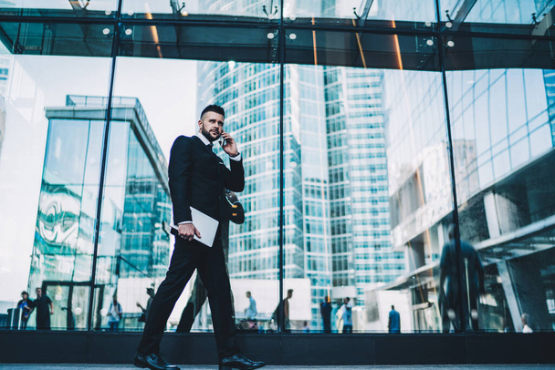 Serious male entrepreneur in formal wear holding laptop in hand and making cellphone conversation during walking in downtown,Caucasian businessman going to enterprise office using roaming for calling - Fotoğraf, Görsel