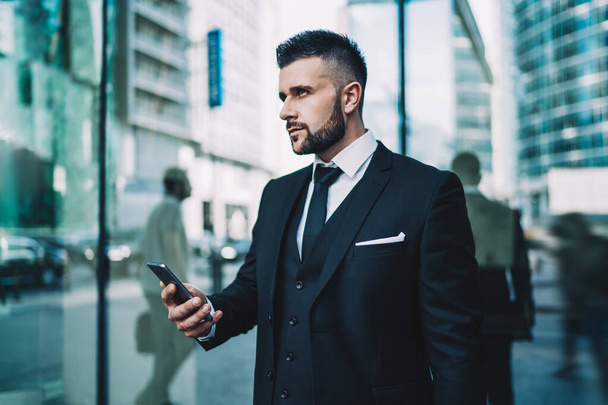 Pensive young man in elegant wear thoughtful looking away and thinking on content from received cell email, Caucasian intelligent trader waiting for personal lawyer in downtown holding smartphone - Foto, imagen