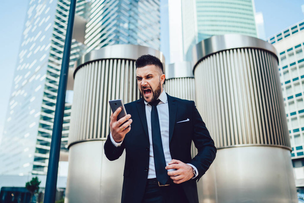 Crazy male entrepreneur feeling anger from trouble with solving enterprise problems shouting on cell smartphone gadget in financial district in megalopolis, unhappy man screaming to mobile gadget - Photo, image