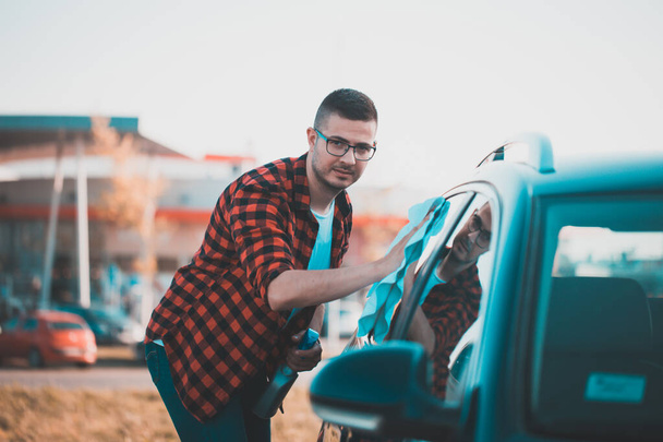 Porträt eines zufriedenen jungen Mannes, der sein eigenes Auto mit einem Tuch reinigt. - Foto, Bild