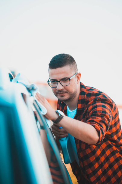 Portrét spokojeného mladého muže, který si čistí auto mikrovláknitou látkou a sprejem v láhvi. - Fotografie, Obrázek