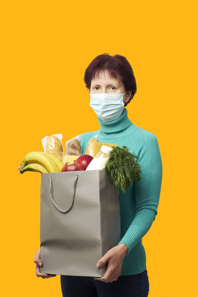 Mulher em uma máscara médica segura um saco de compras de papel com alimentos isolados em fundo laranja
 - Foto, Imagem