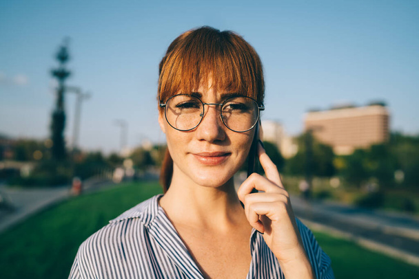 Close up portrait of attractive female millennial in classic glasses for provide eyes protection looking at camera while making cellphone call via application, beautiful woman phoning to operator - Foto, afbeelding