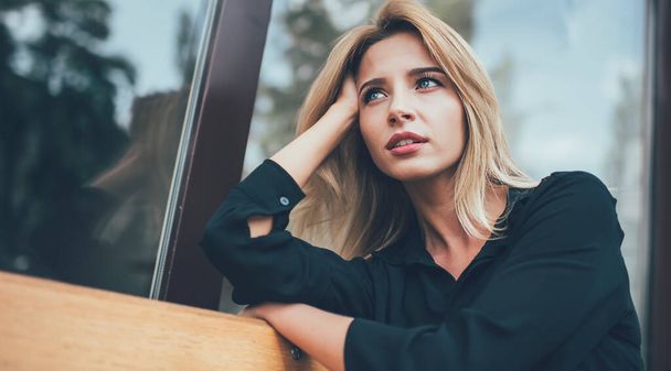 Pensive contemplative hipster girl with gorgeous blonde hair looking away and thinking about spending leisure, attractive female teenager dressed in casual black shirt taking rest on city bench - Foto, afbeelding