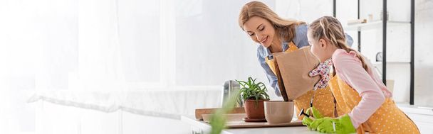 Horizontal image of happy mother with paper bag putting ground to flowerpot and cute daughter with rake near table in kitchen - Photo, Image