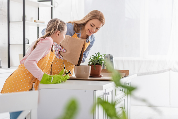 Valikoiva painopiste äiti hymyilee ja laskemisesta maahan kukkaruukku ja tytär puutarhanhoito työkaluja lähellä pöytää keittiössä
 - Valokuva, kuva