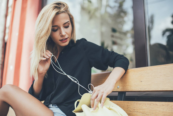 Pretty hipster girl in casual wear getting out electronic headphones from backpack for listening music on urban setting, attractive female meloman searching earphones while taking rest on bench - Photo, Image
