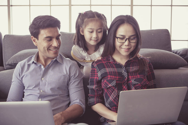 多様な家庭の3人のメンバー、白人の父とアジアの母と小さな半分の娘は、家のリビングルームとノートパソコンを使用して一緒に座っています。自宅で働くためのアイデア. - 写真・画像
