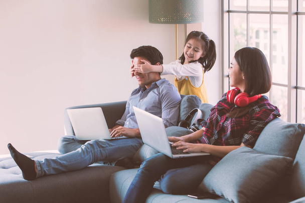 Członkowie zróżnicowanej rodziny, ojciec kaukaski i matka Azji za pomocą notebooka laptopa do pracy z domu, młoda córeczka cute przyjść do żartu jej tata zamykając oczy rękami. - Zdjęcie, obraz