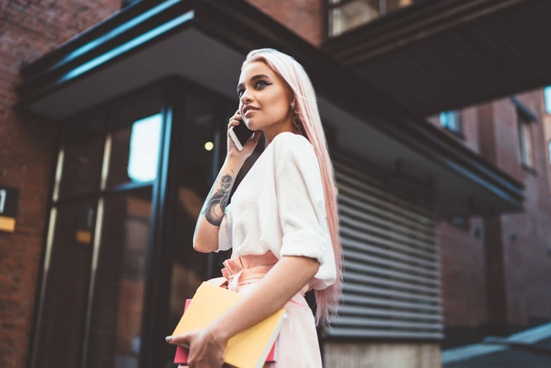 Below view of attractive trendy female teenager with pink hair consultancy with colleague during mobile talking, young hipster girl with tattoo on hand using modern smartphone gadget for phoning - Valokuva, kuva
