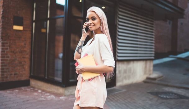 Half length portrait of young stylish hipster girl with long pink hair standing outdoors and making international conversation via cellphone, happy cheerful female student with knowledge notepad - Foto, immagini