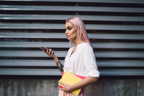 Side view of fashionable female schooler 20s standing on publicity area with notepads for education and smartphone device in hands, stylish hipster girl with colorful textbooks on urban setting - Foto, Imagem