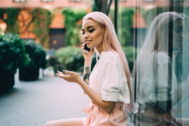 Adolescente femenina positiva vestida con ropa casual descansando en la calle de la ciudad haciendo una llamada internacional al servicio al cliente, atractiva chica hipster milenaria con teléfono celular de pelo rosa
 - Foto, Imagen