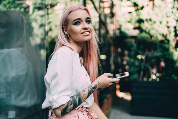 Positive hipster girl with cellular phone in hand looking away while sitting outdoors, stylish teenage woman with youth tattoo on hand holding smartphone device and resting on urban setting - Photo, image