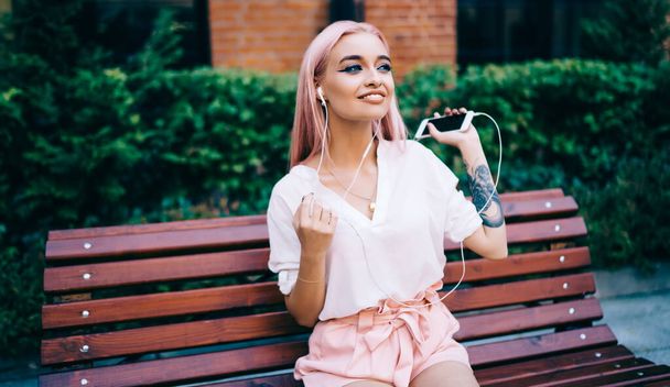 Cheerful woman with beautiful pink hair holding cellphone gadget in hand smiling and looking away during leisure with favourite music,positive female teenager using modern technology for melomany time - Photo, image