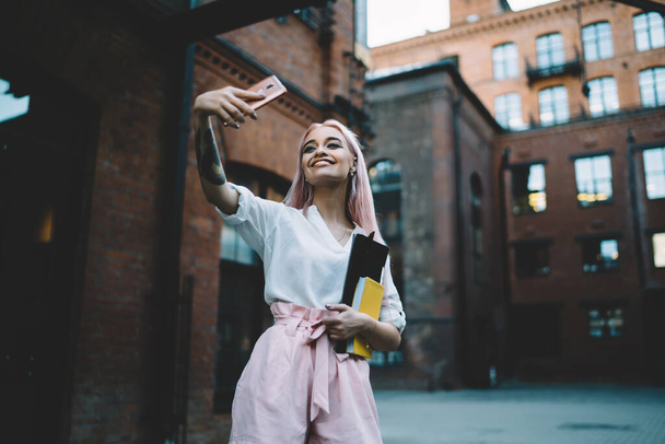 Positive woman holding cellphone for photographing herself via front camera standing on city street, happy cheerful hipster girl clicking images on smartphone for creating publication on website - Фото, зображення