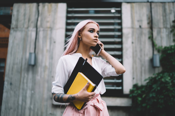 Serious attractive female university student having mobile phone conversation standingg outdoors at city street, beautiful stylish hipster girl 20s holding textbook and phoning via smartphone - Foto, immagini
