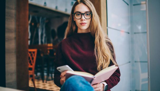 Portrait of serious female reader in optical eyeglasses for provide eyes protection holding textbook for education, young woman with notepad and smartphone gadget in hands sitting in coffee shop - Fotoğraf, Görsel