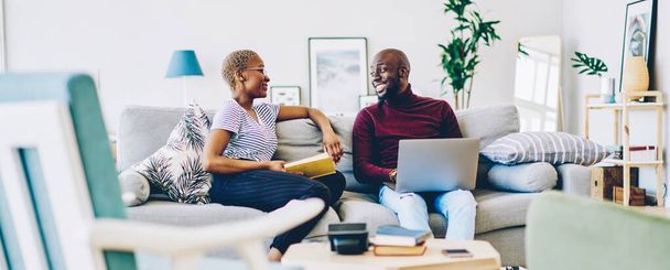Cheerful african american marriage spending time at home enjoying free time together, positive woman sitting with book while communicating with her husband using laptop computer and wifi in apartmen - Foto, Bild