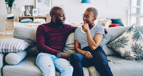 Young african american marriage  having conversation spending time at home interior together, Handsome dark skinned man looking at his girlfriend hugging sitting on cozy sofa in modern living roo - Foto, Imagem