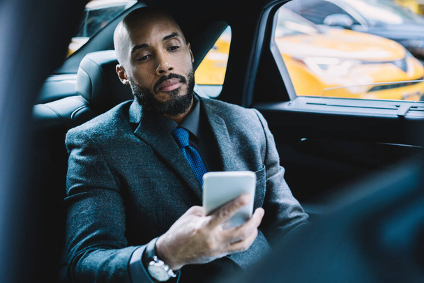 African American businessman using digital technology for development project during transportation in comfortable luxury car to office building, serious male expert sending sms with information - Photo, image