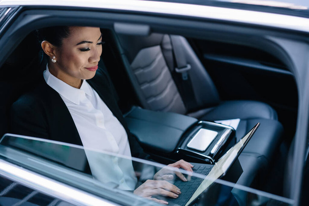 Erfolgreiche Bankerin in festlichem Gewand sitzt auf dem Rücksitz des Autos und tippt auf einer tragbaren Tastatur, die mit einem digitalen Tablet verbunden ist, mittels 4g-WLAN während der Geschäftsreise den Vertrag ein - Foto, Bild