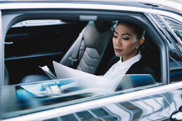 Attractive businesswoman reading details from companies contract while getting to office destination on backseat of luxury car, clever female expert concentrated on information from accounting - Foto, immagini