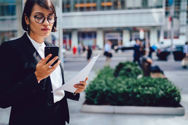 Concentrated businesswoman dressed in formal wear and eyeglasses standing on New York street with documents and modern smartphone device and checking accounting on financial website and papers - Zdjęcie, obraz