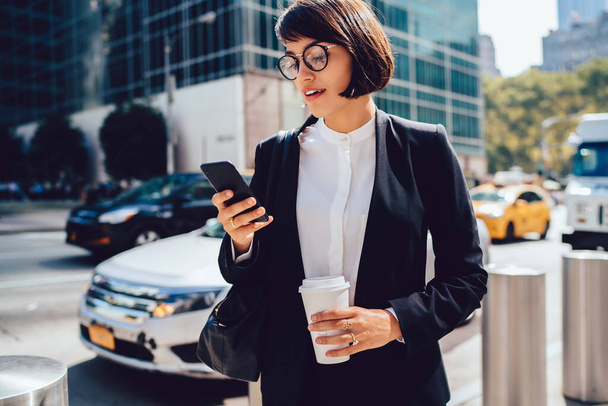 Attractive female owner of bank reading incoming message on smartphone while standing on street near traffic, brunette female employee chatting online with business partners during work break - Foto, imagen