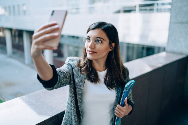 Attractive millennial hipster girl in stylish eyewear clicking selfie photos via front camera on modern cellular gadget, beautiful woman with textbook for education taking pictures via app - Photo, Image