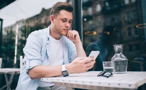 Handsome pensive millennial man browsing mobile phone while relaxing at white table near window on outside terrace of city cafe - Fotografie, Obrázek