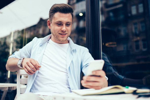 Contemporary millennial man surfing mobile phone while sitting relaxed at table on street terrace of cafe and smiling in leisure - Valokuva, kuva