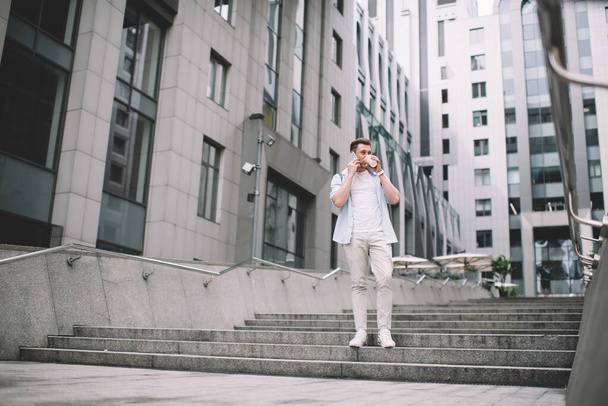 Modern millennial man drinking coffee and speaking on mobile phone while standing on street steps with modern city buildings on background - Foto, immagini