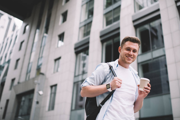 From below of handsome millennial man with backpack having cup of coffee to go while smiling at camera on street - Photo, image
