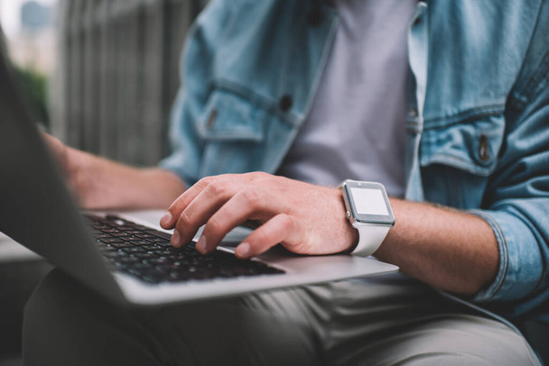 Faceless modern millennial man wearing casual outfit and smart watch with white screen sitting on street and working on contemporary laptop  - Foto, imagen