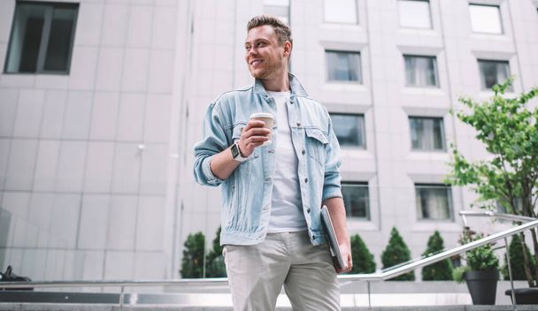 From below of handsome millennial man in casual outfit having cup of fresh coffee and standing on street with laptop smiling away - Foto, afbeelding