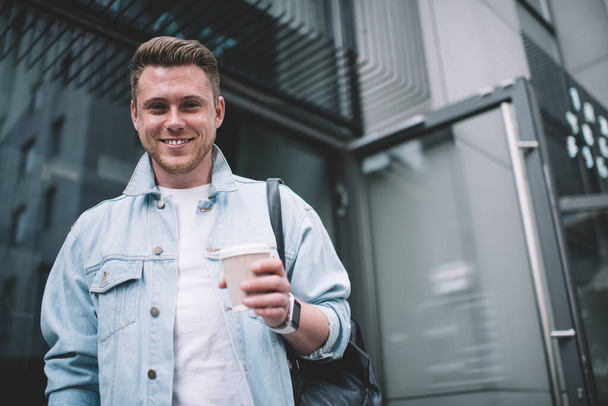 From below of smiling young Caucasian man in stylish denim jacket and with backpack holding cup of coffee looking at camera - Photo, image