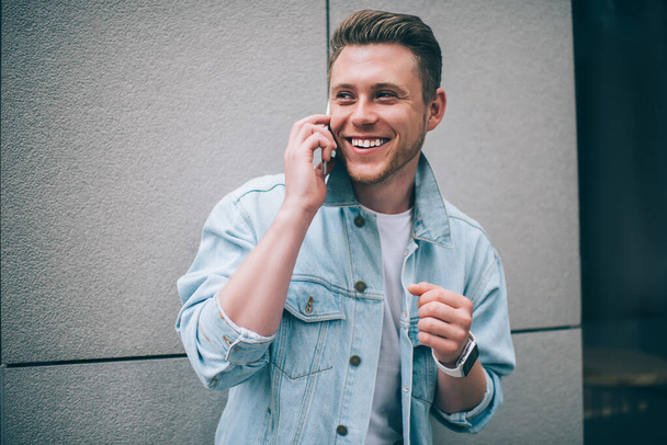 Handsome millennial man in cool denim jacket laughing happily while looking away and chatting on mobile phone standing on street - Фото, изображение