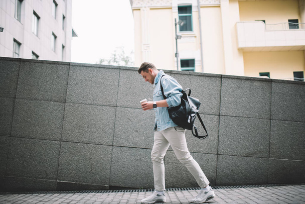 Вид сбоку трогательного случайного человека в джинсовой куртке и с черным рюкзаком, держащим чашку кофе, идущего по улице
 - Фото, изображение