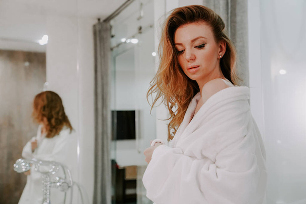Een mooie vrouw na een douche in een witte badjas kijkt in de spiegel in het bad. Zelfgemaakte lichaamsverzorging. spa behandelingen - Foto, afbeelding