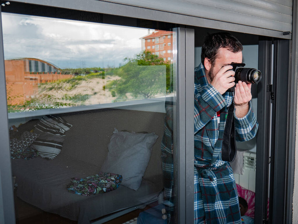 Homme mal aligné en peignoir à carreaux prenant des photos de sa terrasse à la maison caché sous l'aveugle pendant le confinement en raison de la vigilance coronavirus
 - Photo, image